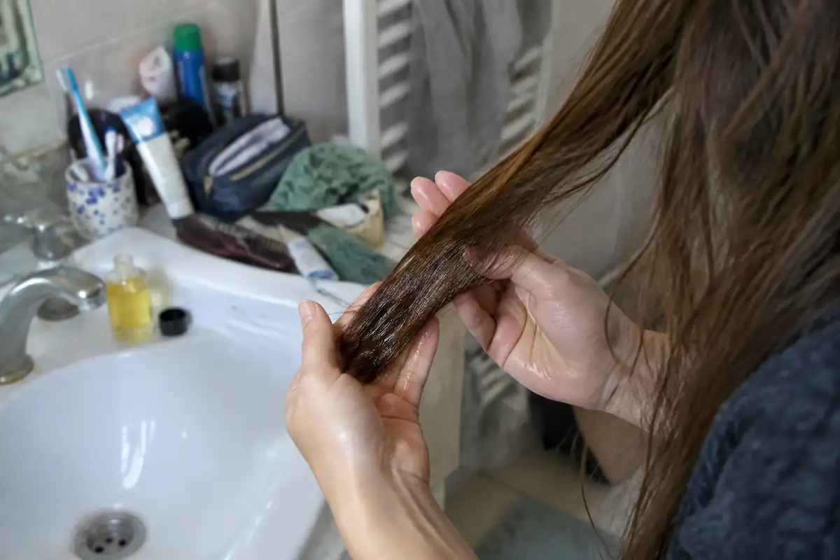Mains répartissant une petite quantité d'huile sur une mèche de cheveux secs au-dessus d'un lavabo
