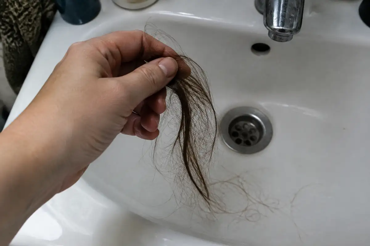 Une main tient une mèche de cheveux tombés au-dessus d'un lavabo
